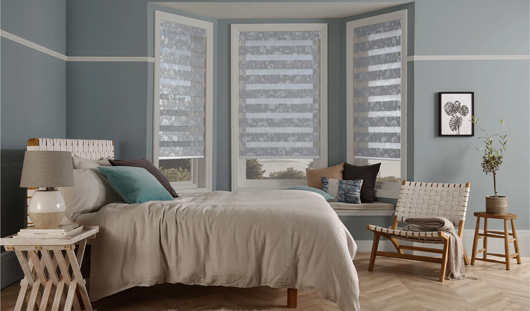 Modern bedroom with gray walls, bay windows with patterned blinds, beige bed, chair, and wood floors.