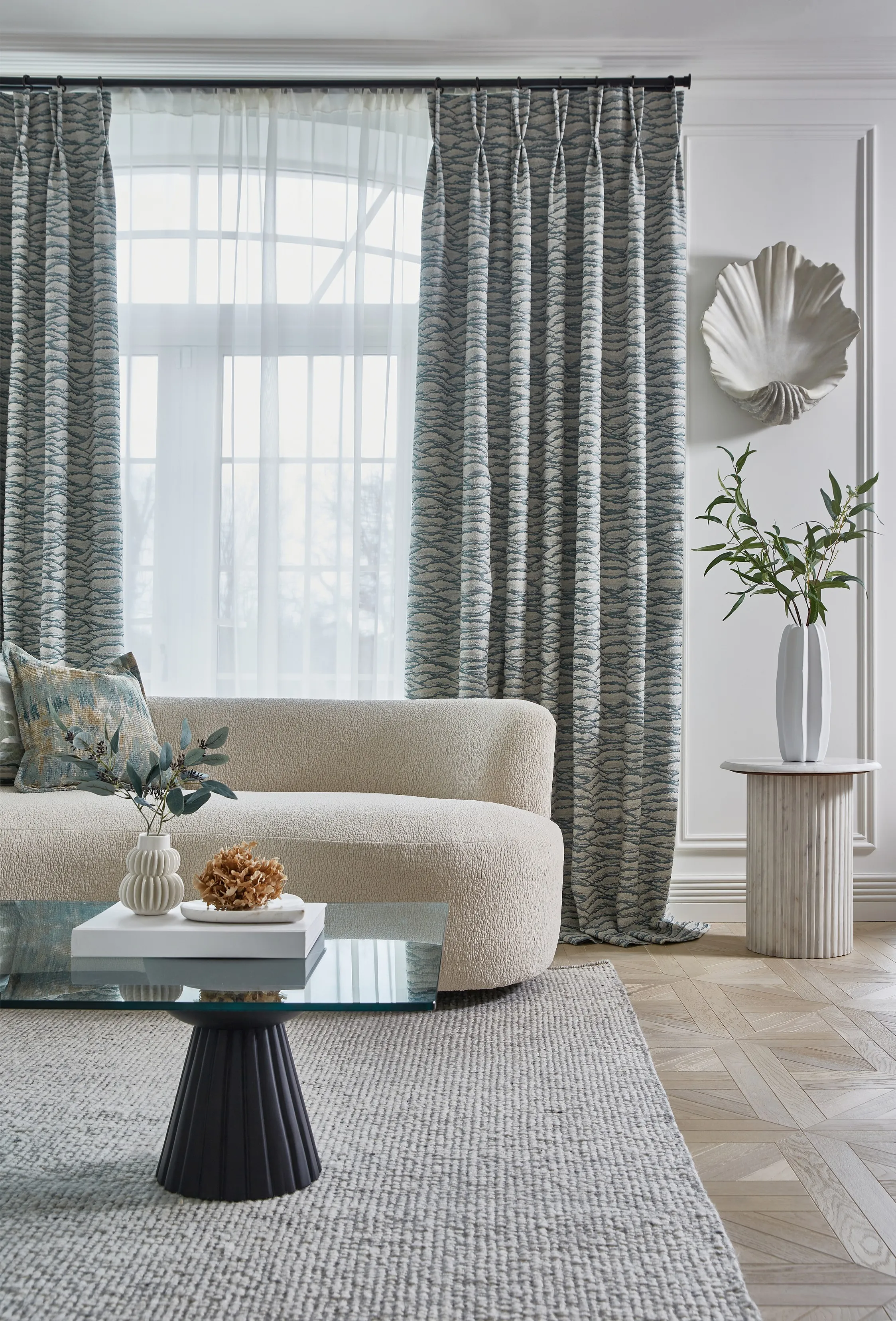 Modern living room with textured beige sofa, patterned curtains, glass coffee table, and decorative plants.