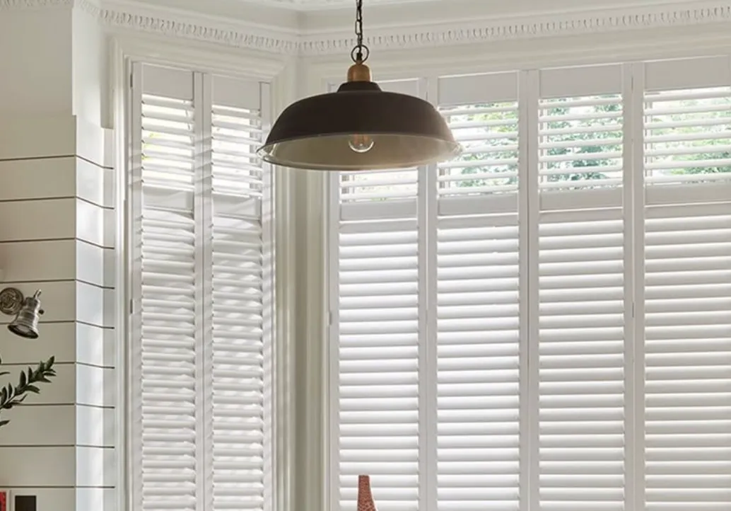 White window shutters covering large windows with a black hanging pendant light in front.