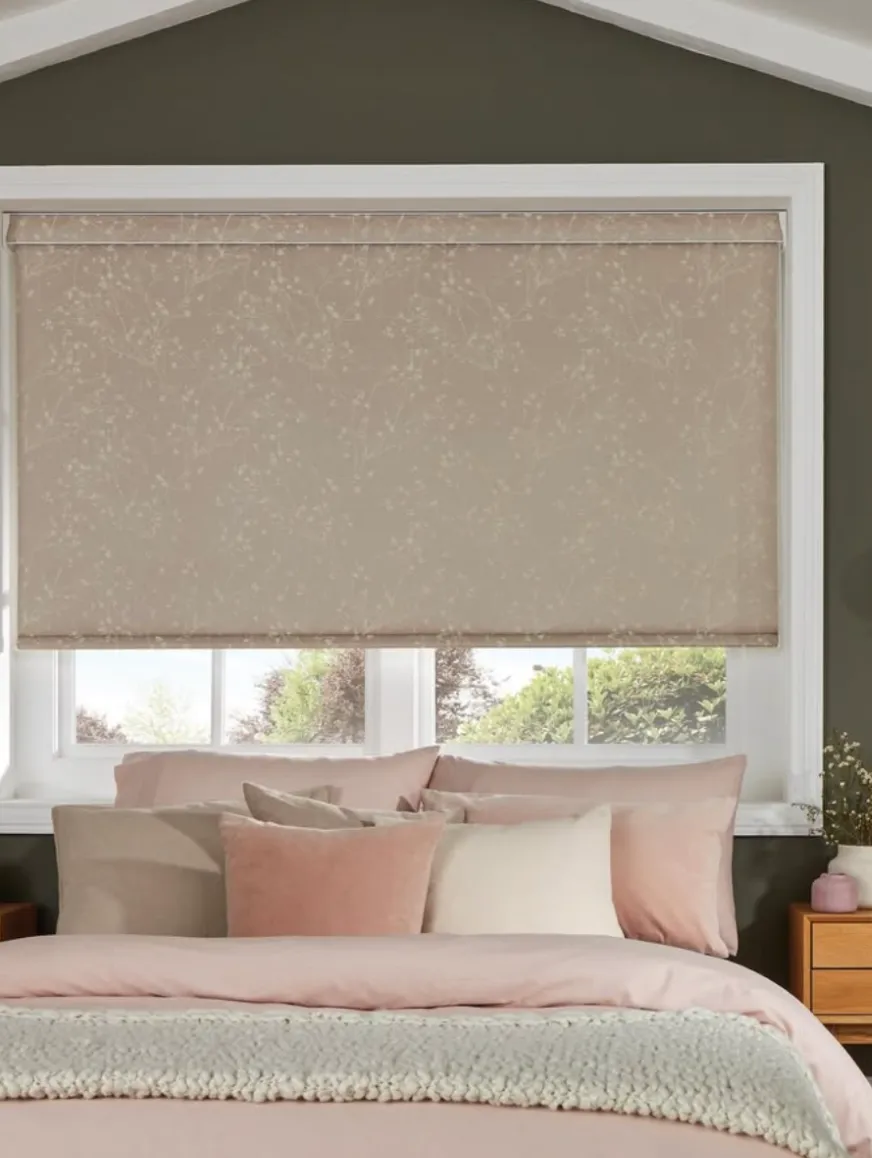 Bedroom window with beige floral roller blind above bed with pink and cream pillows and blanket.