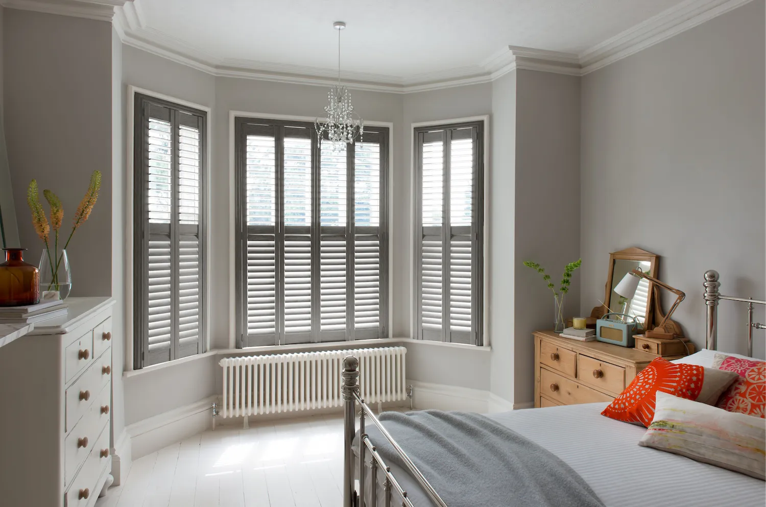 Modern bedroom with gray shutters, metal bed, wooden dresser, and colorful pillows in soft light.