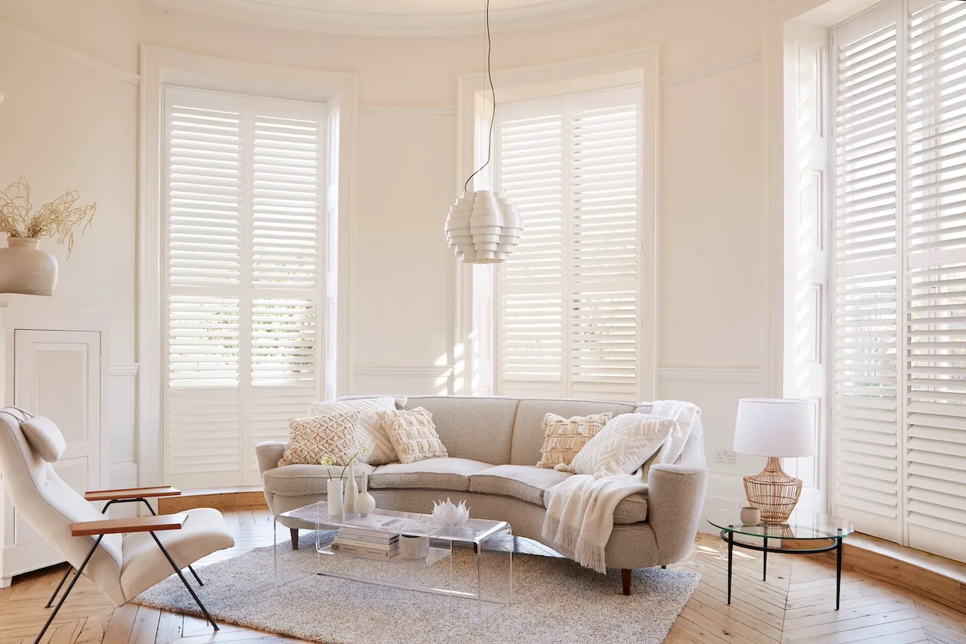 Bright living room with beige curved sofa, armchair, glass tables, and white plantation shutters.