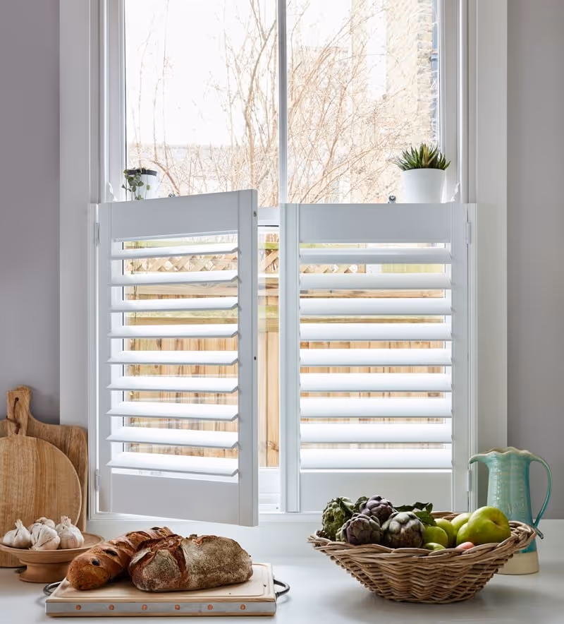 White kitchen window with half-open plantation shutters, bread, garlic, and a fruit basket on the counter.