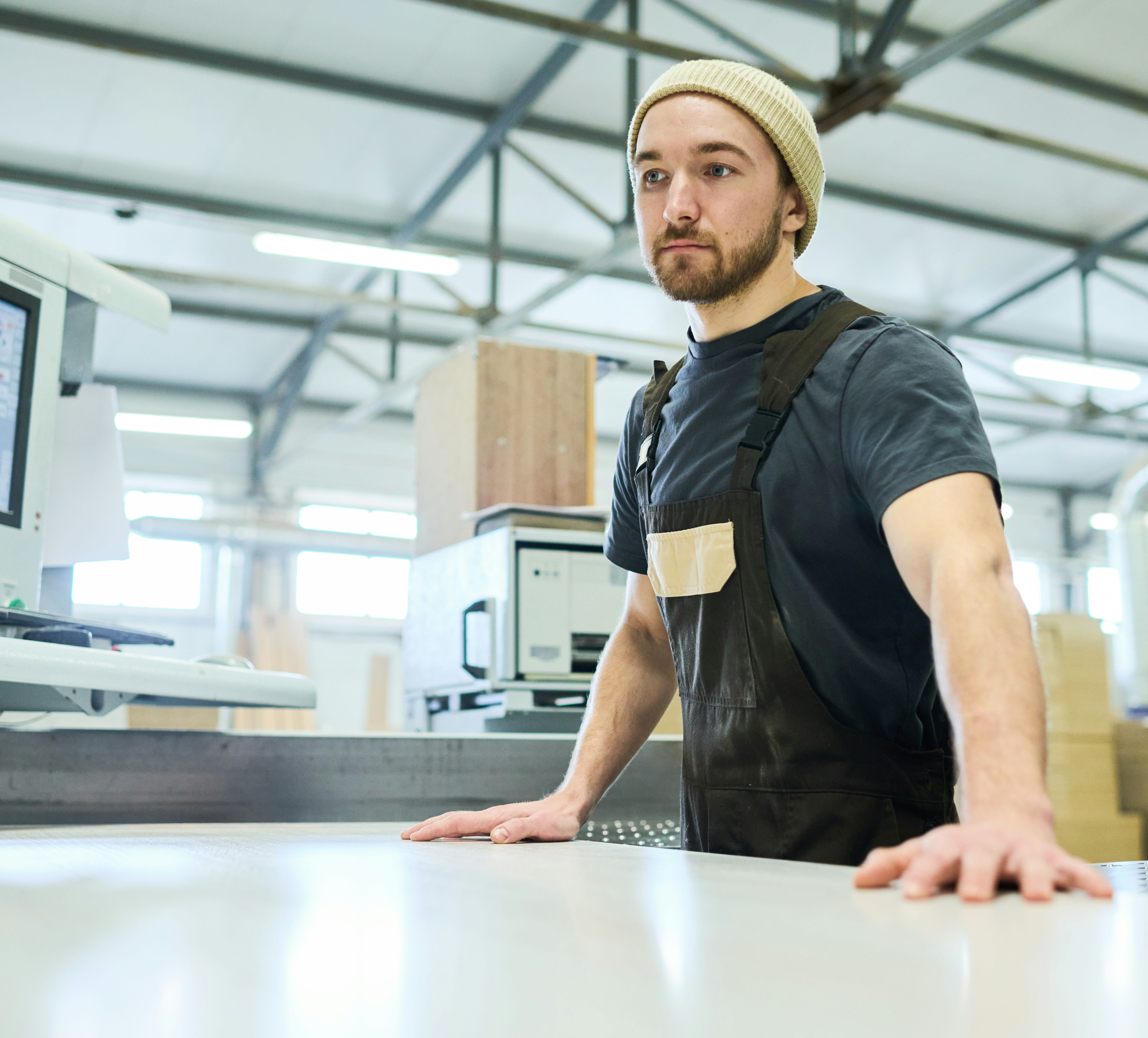 Young factory worker in overalls and beanie standing by a table in an industrial workspace.