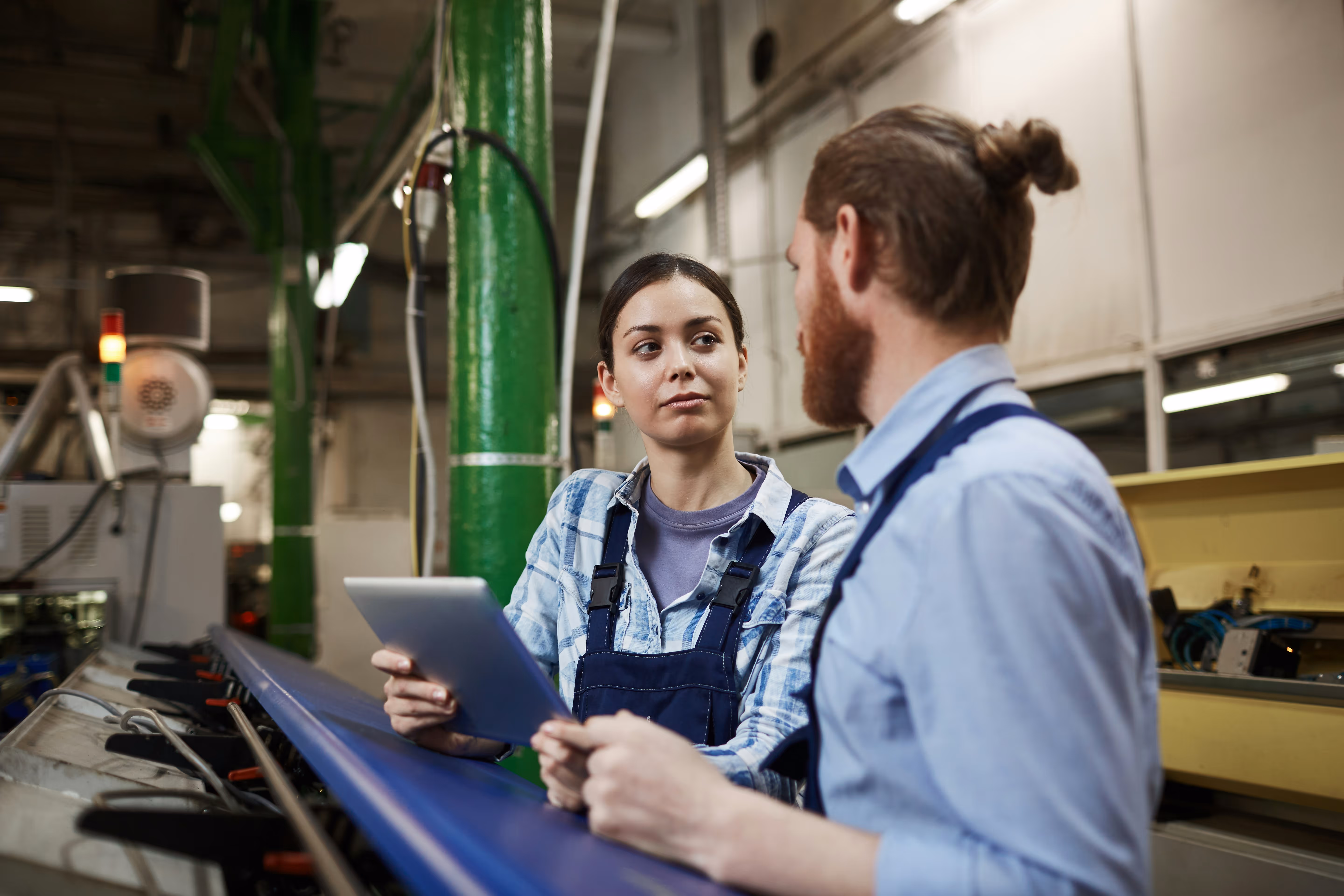Two workers in overalls discussing while one holds a tablet in a manufacturing facility.