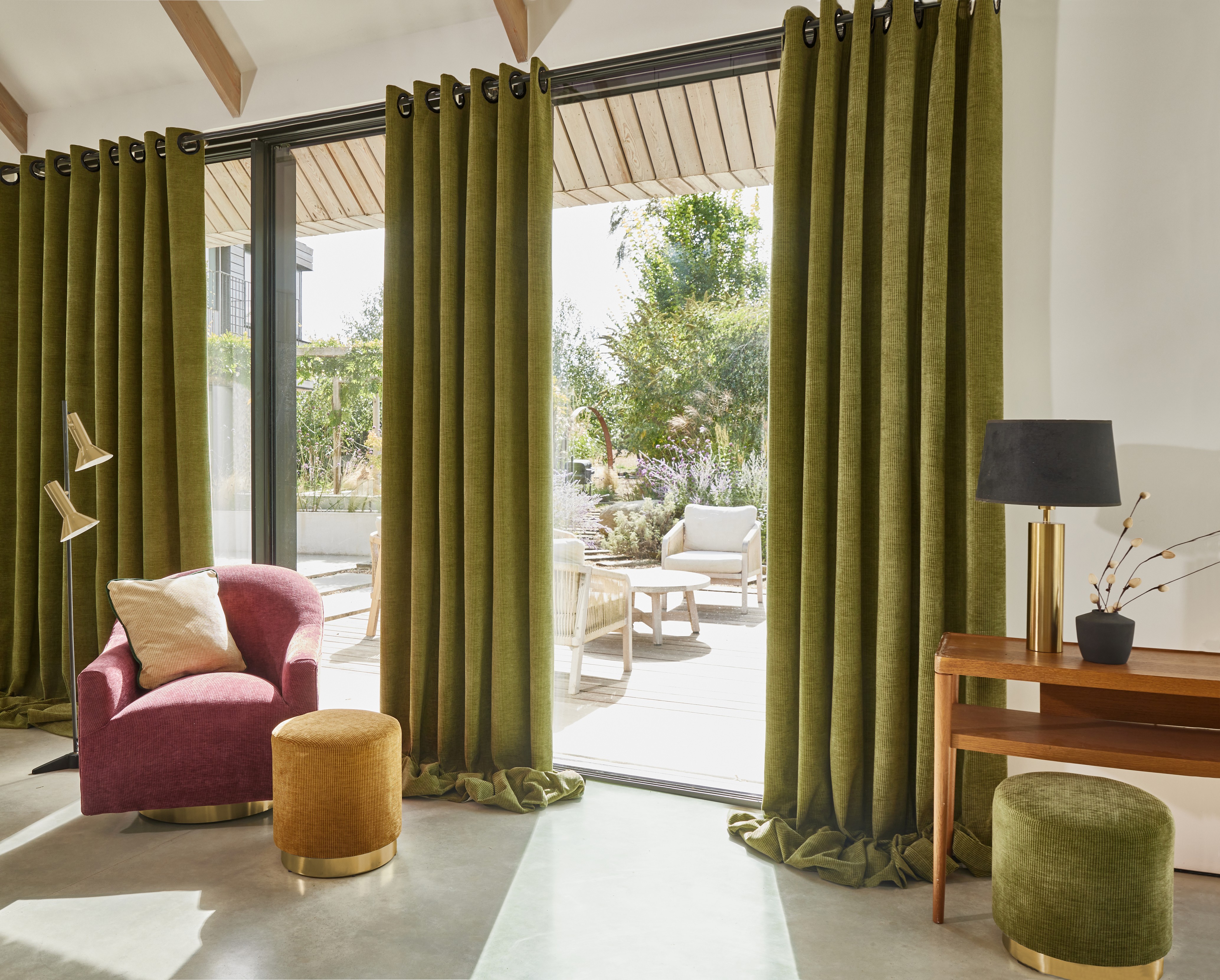 Living room with green curtains, pink armchair, two ottomans, wood table, and outdoor garden view.