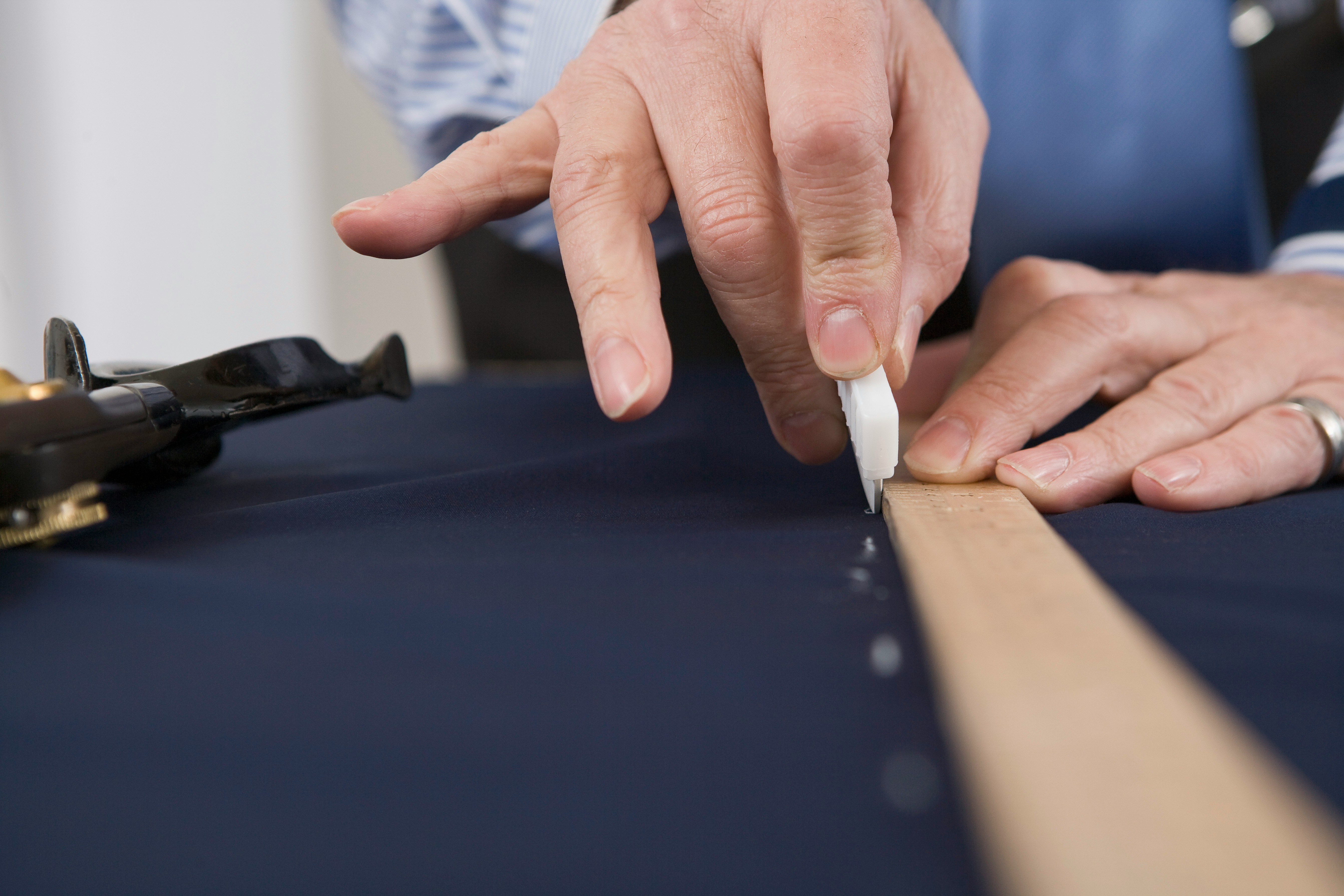 Close-up of hands using a tool to cut fabric along a wooden ruler on a dark surface.