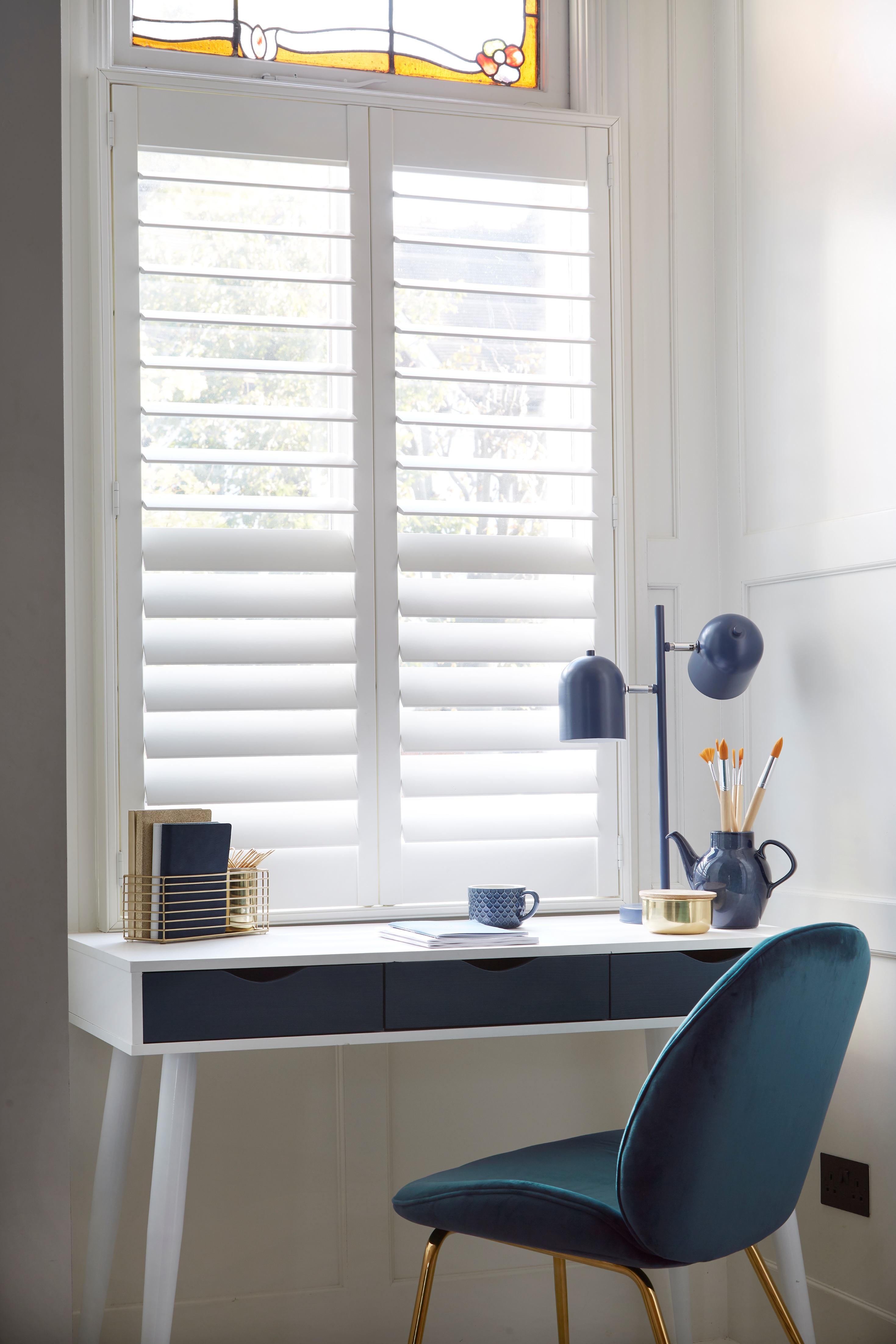 White desk with dark drawers under window shutters with a blue chair and art supplies on top.