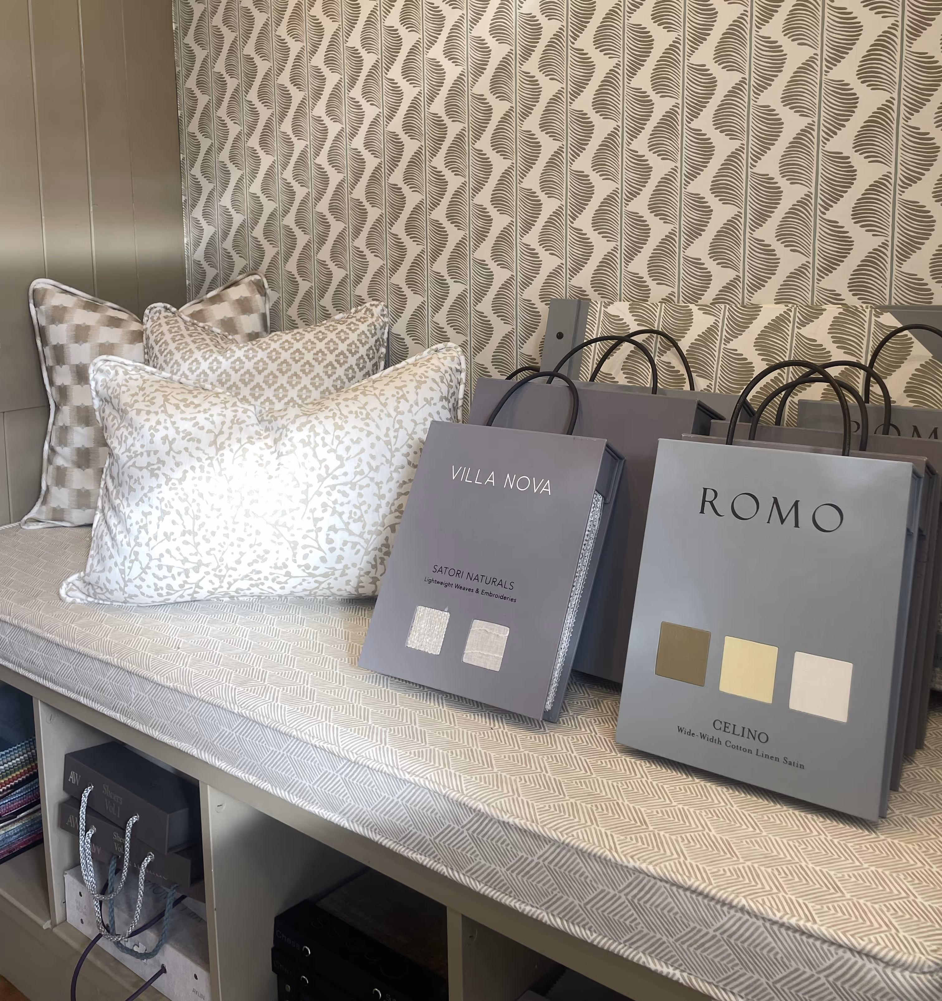 Bench with patterned pillows and fabric sample books labeled VILLA NOVA and ROMO in a showroom.
