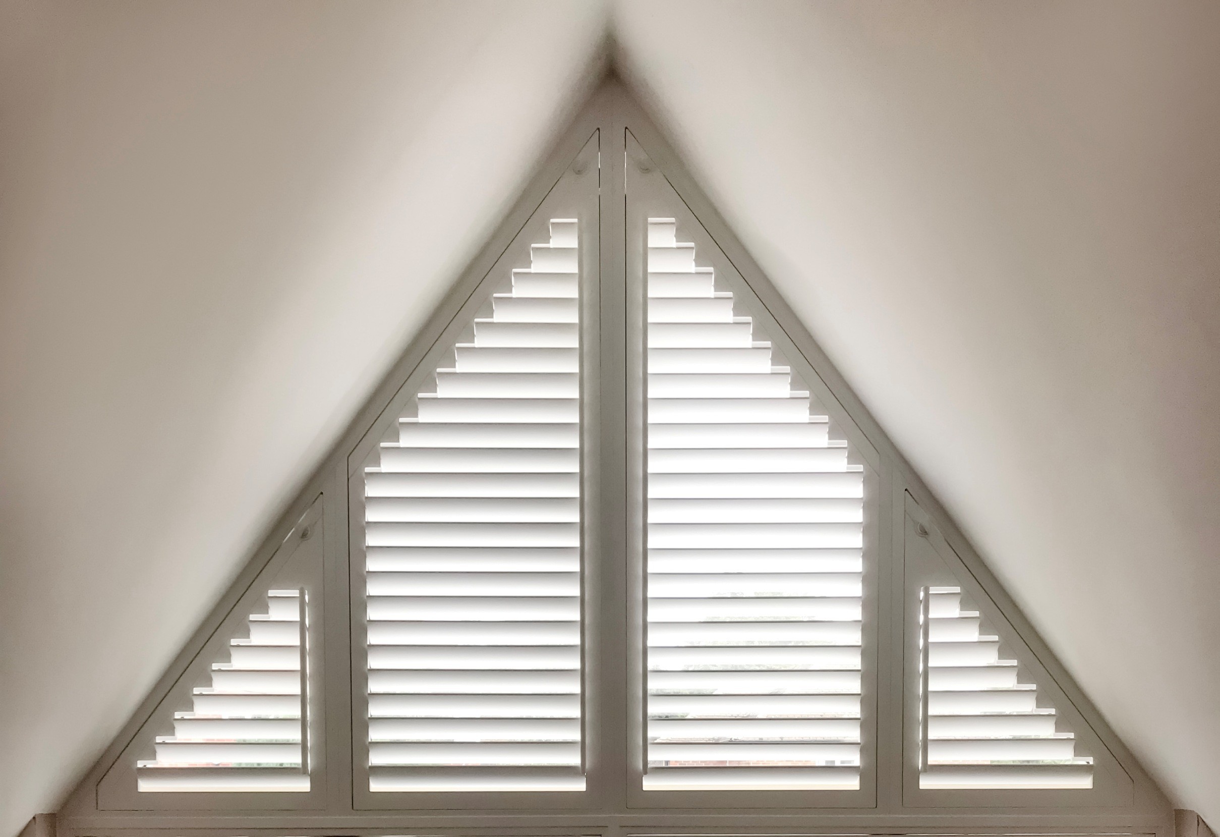 Triangular window with white stepped shutters inside a room with slanted ceiling.