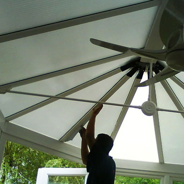 Person installing or adjusting a translucent conservatory roof panel under a ceiling fan.