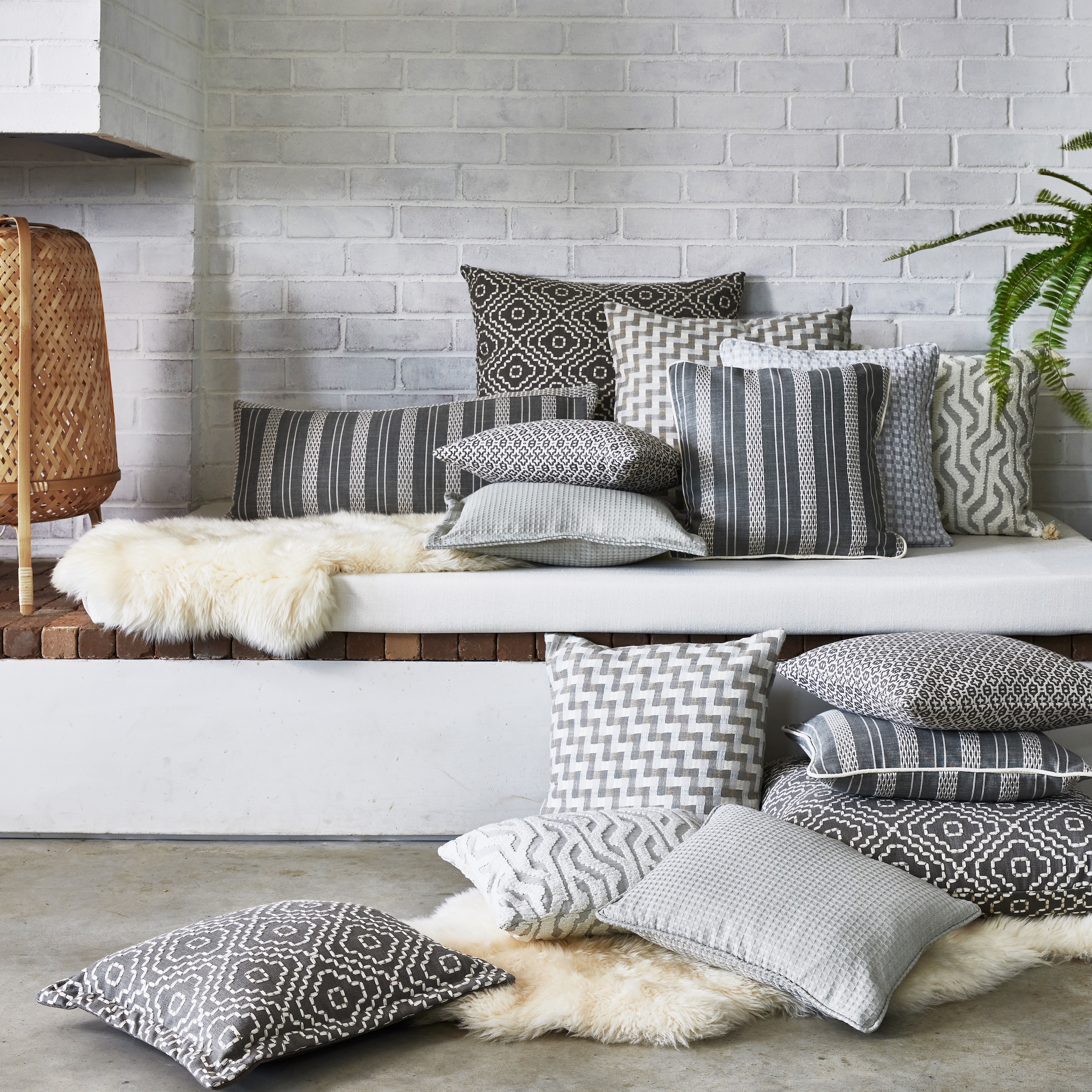 Stacked decorative pillows with geometric patterns on white bench and floor, near a wicker lamp and plant.