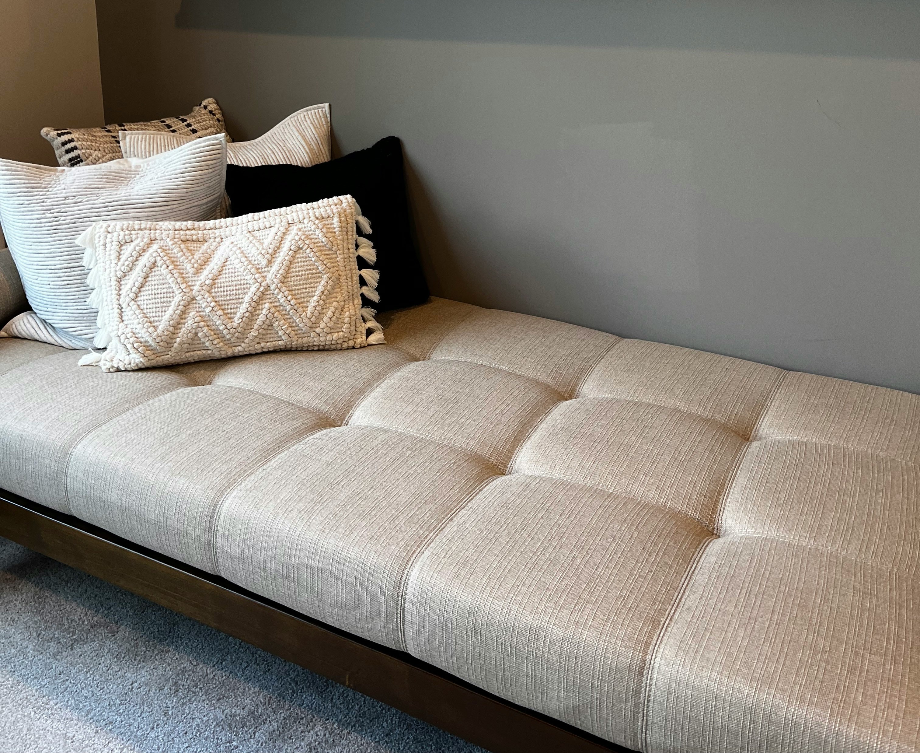 Beige cushioned bench with patterned pillows against a gray wall on a carpeted floor.