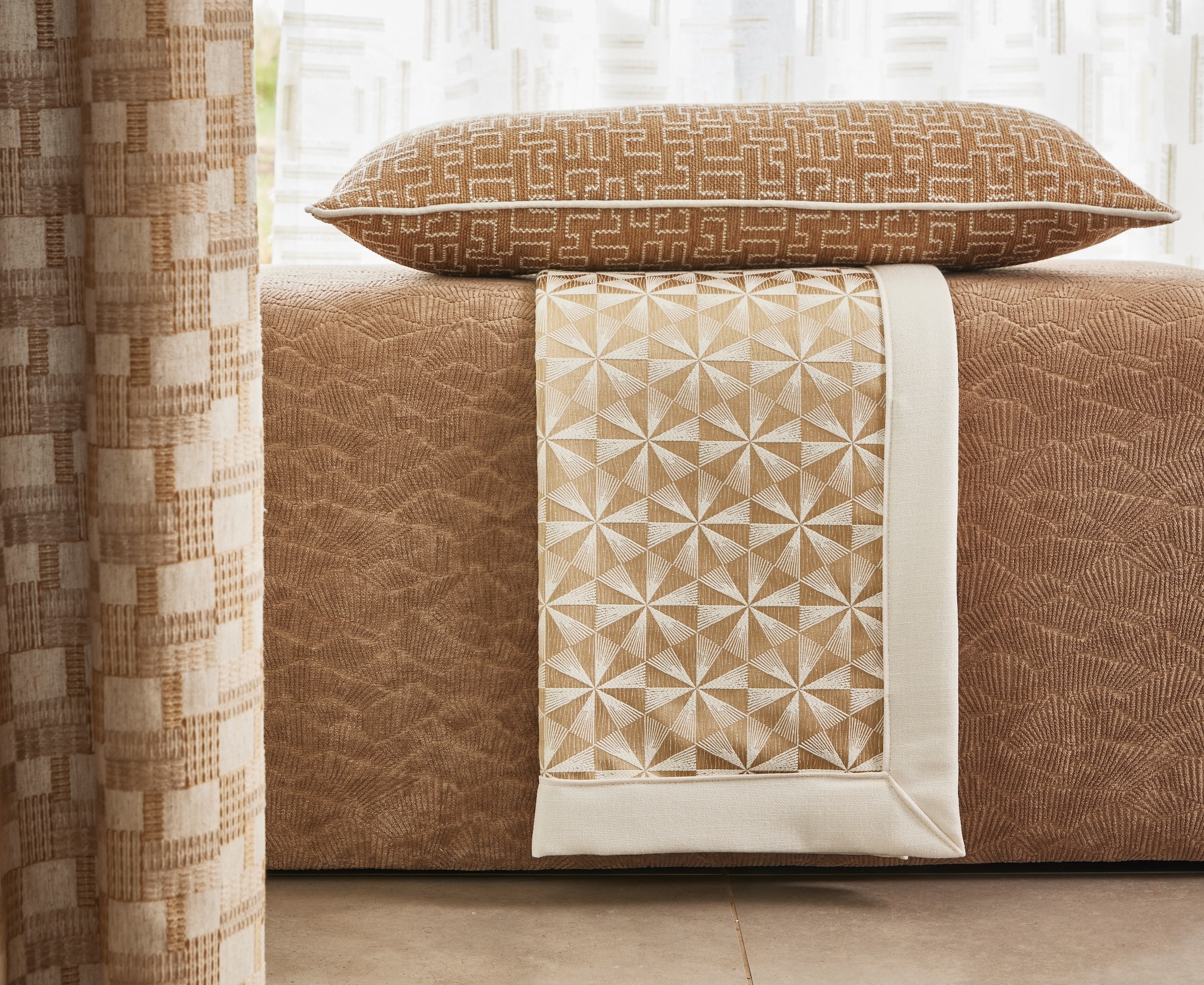 Beige patterned pillow on a textured ottoman with a geometric patterned runner on tiled floor.