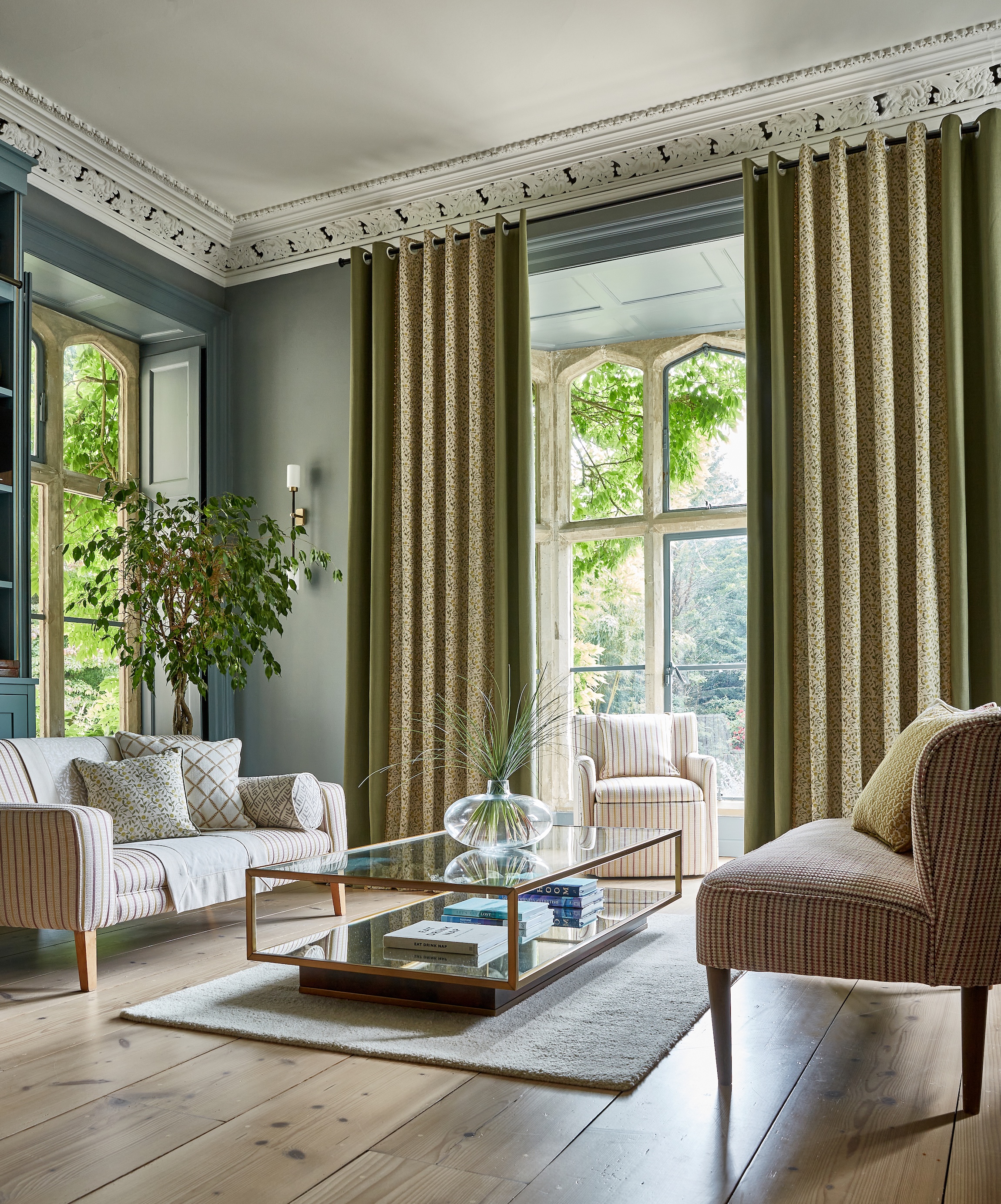 Elegant living room with patterned sofas, large windows, green curtains, glass coffee table, and indoor plant.