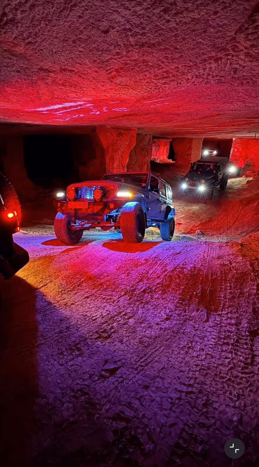 Mehrere Geländewagen fahren in einem Tunnel oder einer Höhle, beleuchtet von roten und violetten Lichtern, mit hellen Scheinwerfern, die den unebenen Boden aus Sand oder Stein erhellen.