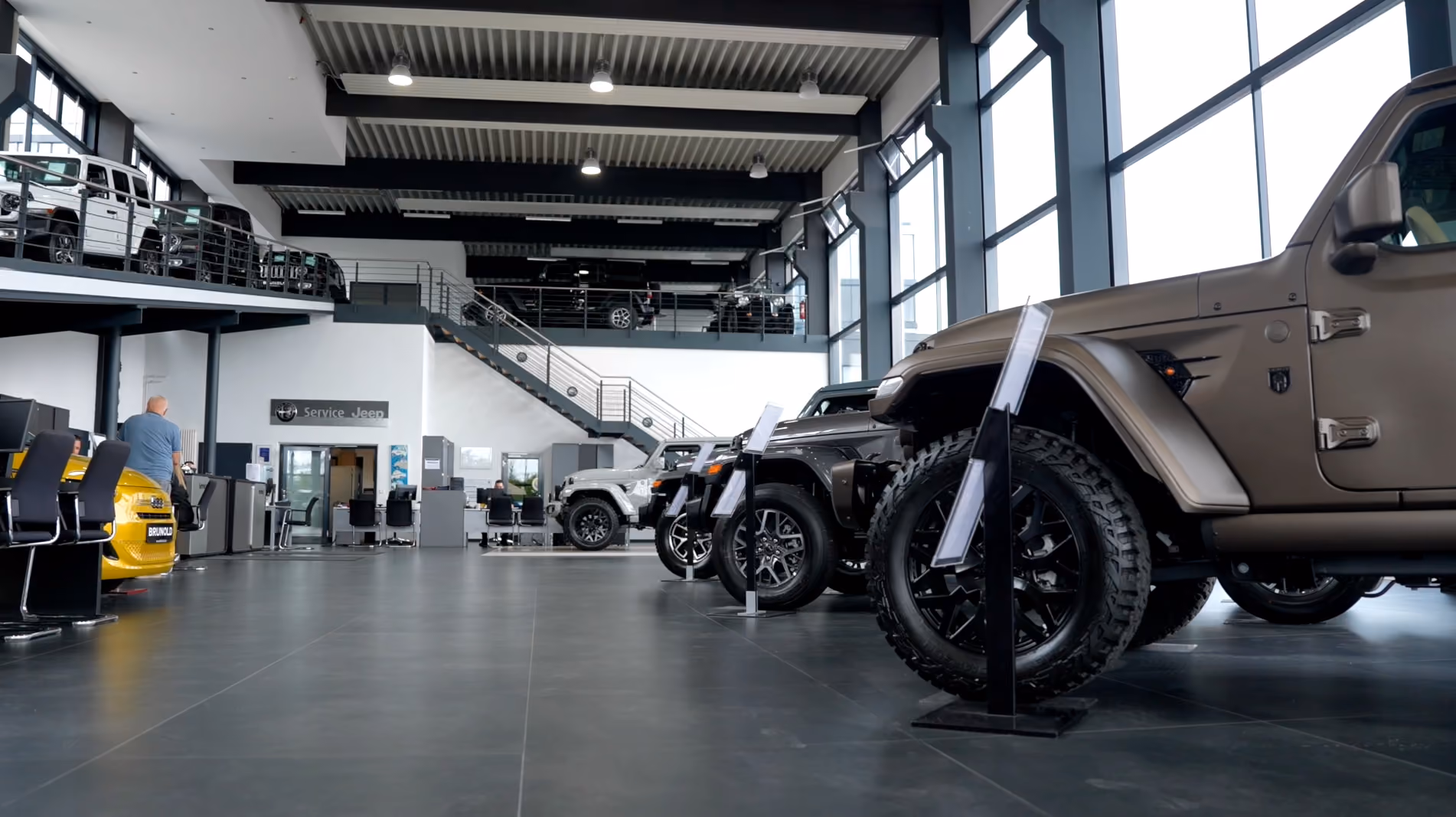 Innenaufnahme eines modernen Autohauses mit mehreren Jeep-Fahrzeugen, die in einer Reihe stehen, große Fenster an der rechten Seite und eine Empore mit weiteren Fahrzeugen im Hintergrund.
