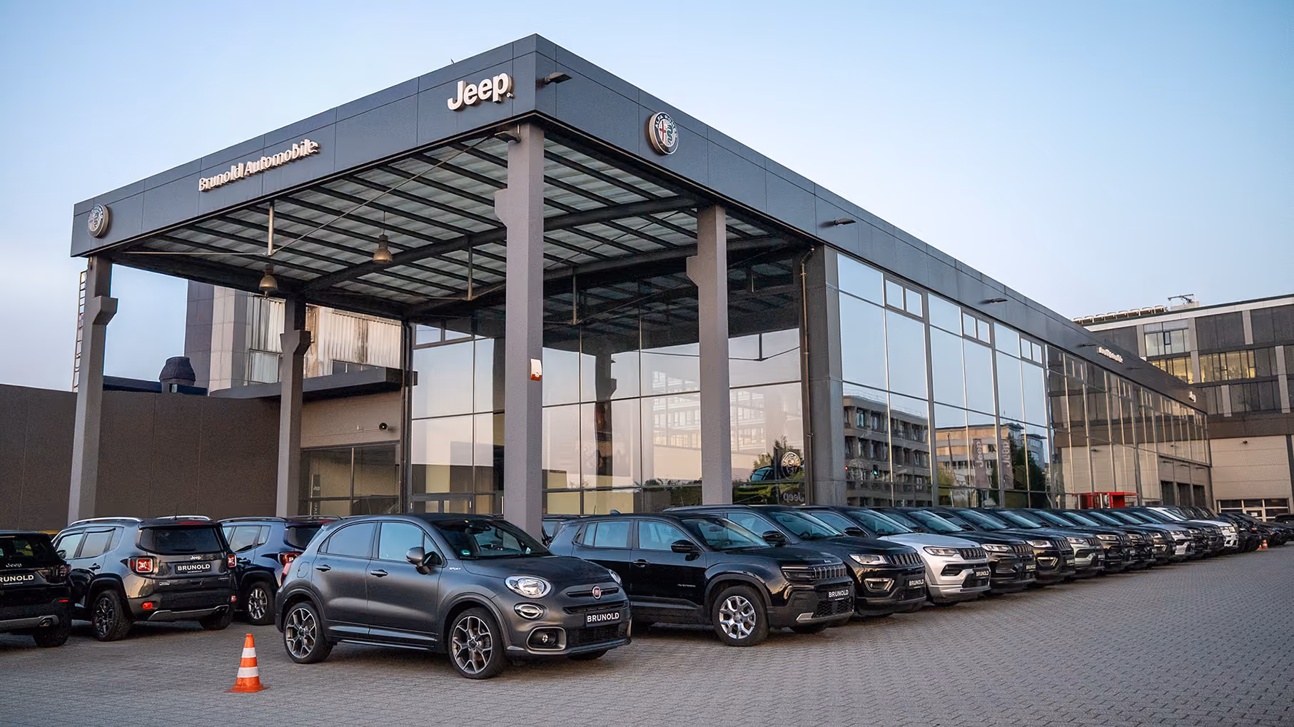 Ein modernes Autohausgebäude mit Glasfronten und vielen vor dem Gebäude geparkten Neuwagen verschiedener schwarzer und weißer Modelle, hauptsächlich SUV-Typen, mit dem Schriftzug ‚Brunold‘ auf den Nummernschildern und Logos von Jeep und Alfa Romeo am Gebäude.