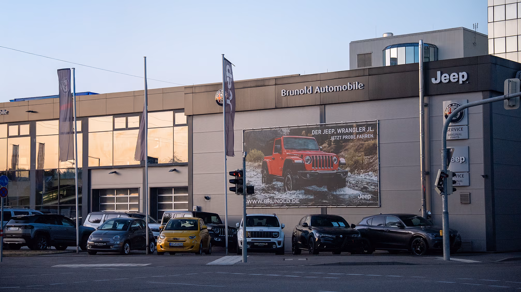 Außenansicht eines Autohauses mit dem Namen Brunold Automobile, das Jeep-Verkauf und Service anbietet, mit mehreren geparkten Autos vor dem Gebäude; ein großes Werbebanner zeigt einen roten Jeep Wrangler, der durch Wasser fährt.