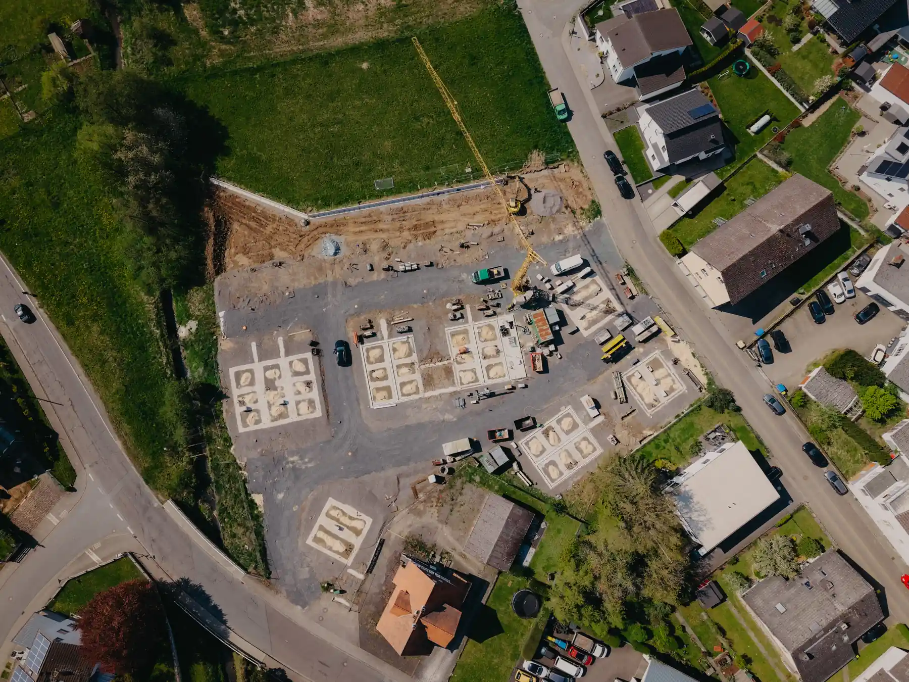 Luftaufnahme einer Baustelle mit Fundamenten, Kran und umliegenden Wohnhäusern.