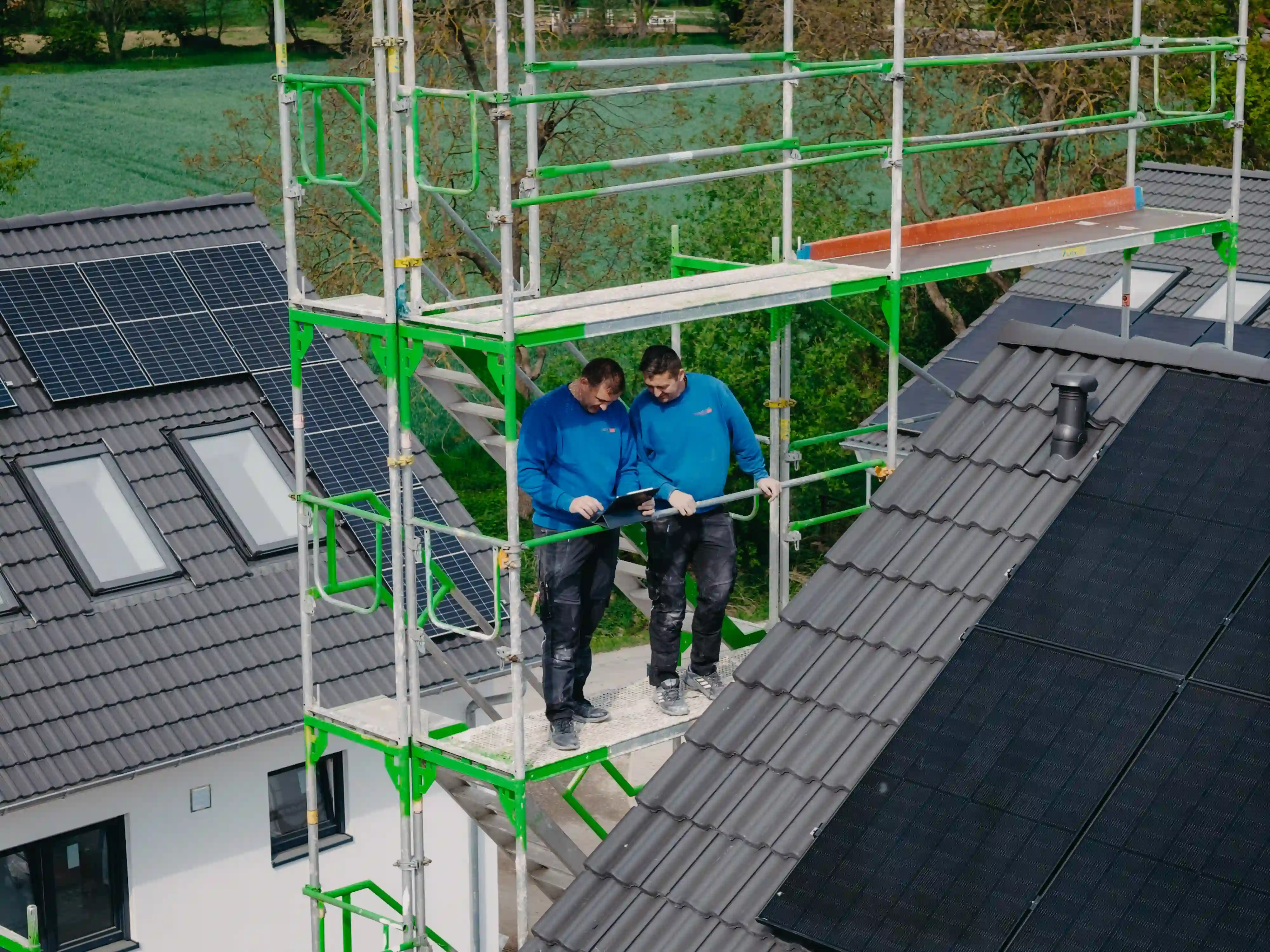 Zwei Arbeiter auf einem Gerüst prüfen ein Tablet neben Solarpanels auf einem Hausdach.