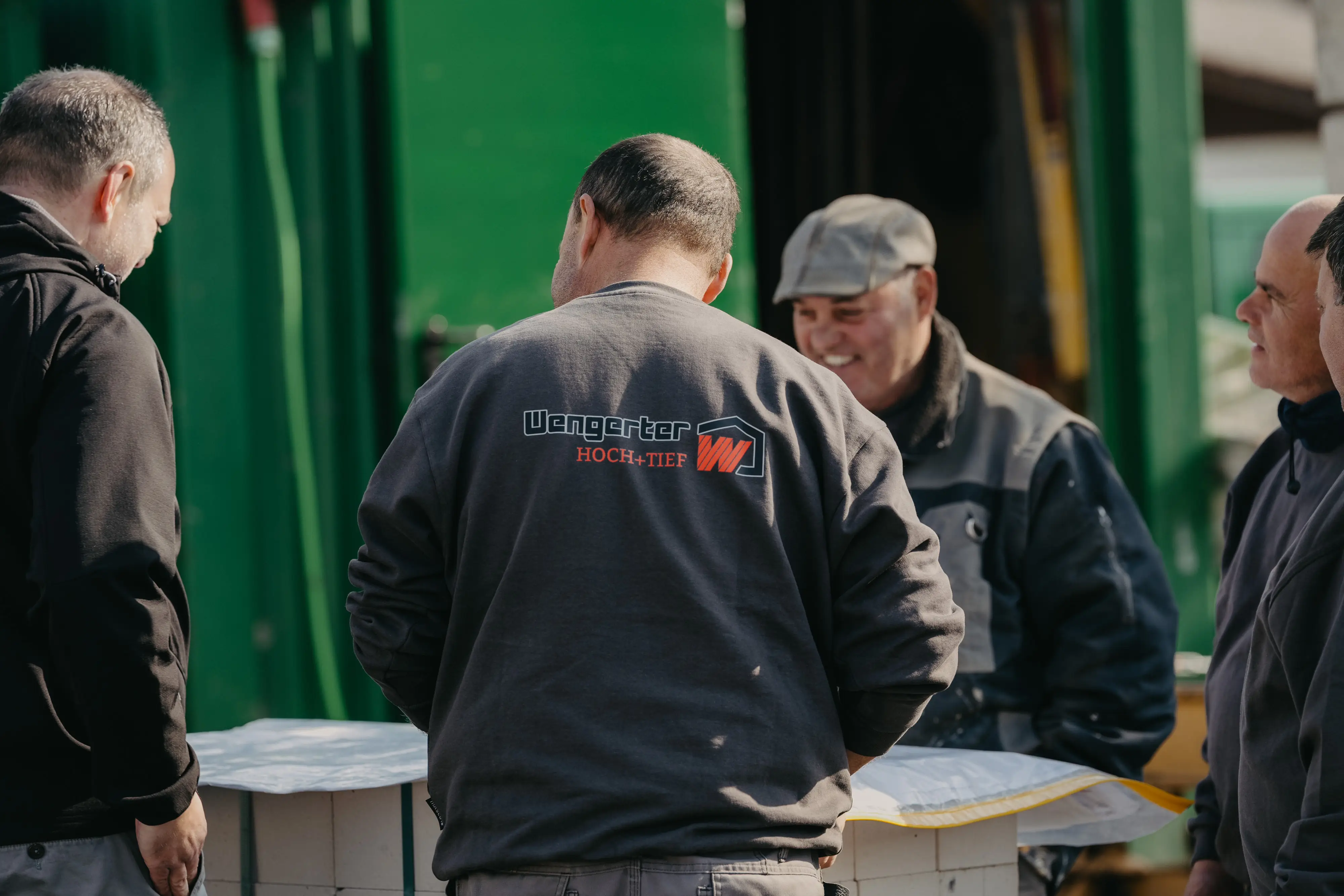 Fünf Männer unterhalten sich in Arbeitskleidung vor grünem Hintergrund, einer trägt ein Hemd mit Wengerter-Logo.