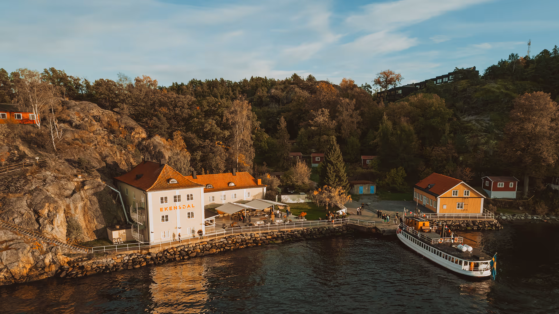 Hus vid vatten med brygga, passagerarbåt och skog i bakgrunden under en blå himmel.