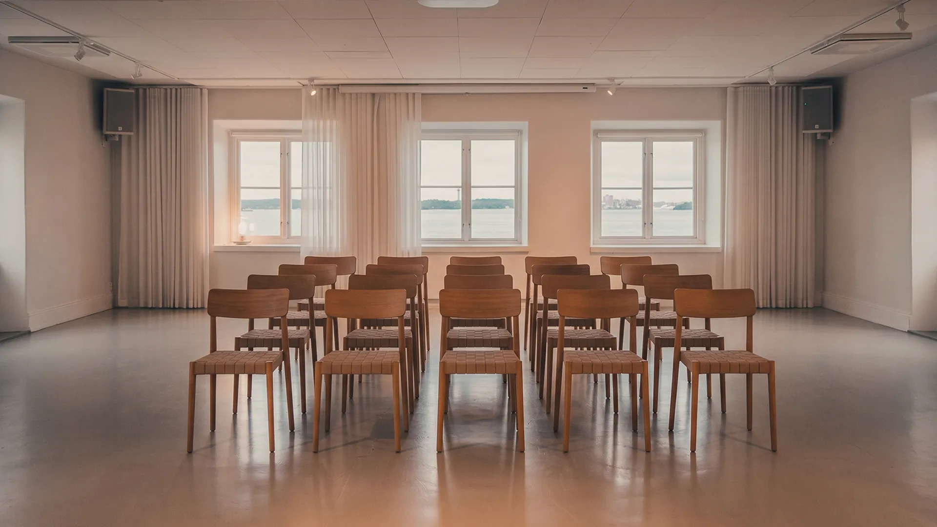 Konferensrum med rader av trästolar och fönster med utsikt över vatten.