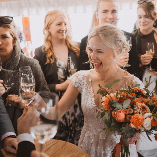 Brud med blomsterbukett ler och skålar med gäster under bröllopsfesten.