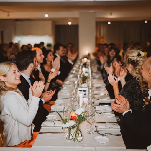 Människor sitter vid ett långbord och applåderar i en festlig middagssal.