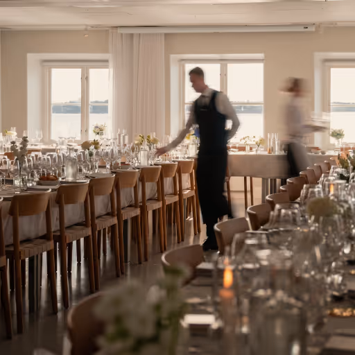 Servitörer förbereder ett elegant dukat långbord med glas och blommor i ett ljust rum.
