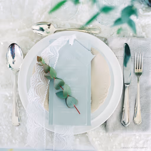 Elegant table setting with white plates, lace napkin, eucalyptus leaf, and silver cutlery on a light tablecloth.