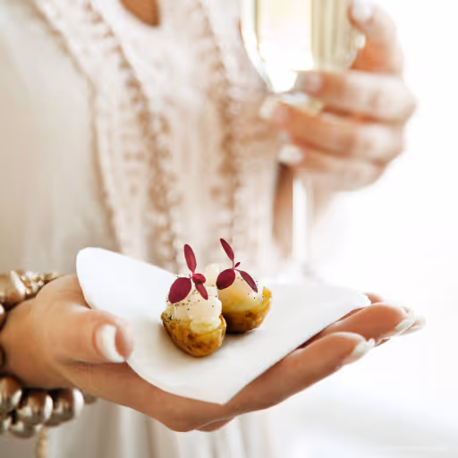 Person håller ett glas champagne och en servett med två små gourmettilltugg med kräm och röda blad.