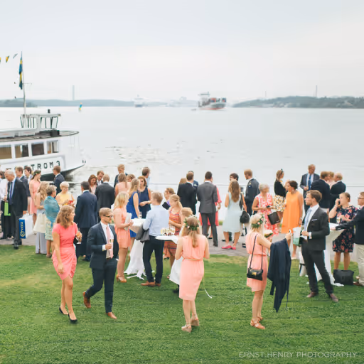 Minglande festdeltagare i sommarkläder vid vattnet nära en båt på gräsmatta.