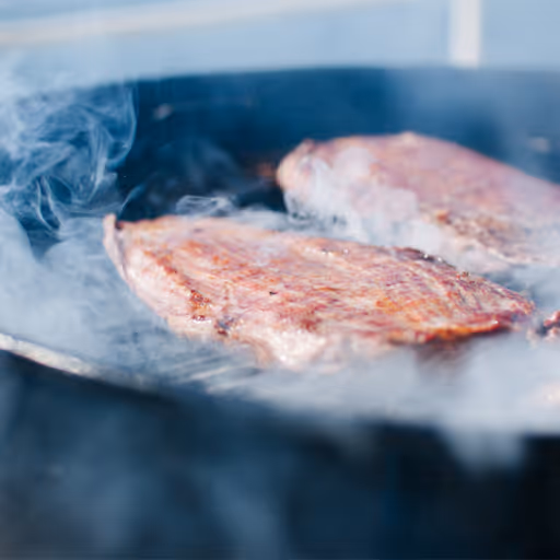 Två köttbitar som grillas på en rökig kolgrill med synlig rök omkring.
