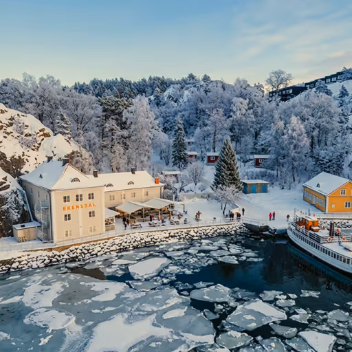 Snötäckt hamn med iskalla vatten, vita byggnader och människor på en brygga i vinterskog.
