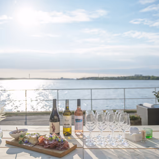 Fyrt glas och flaska vin uppställda på bord utanför med utsikt över vatten och himmel.