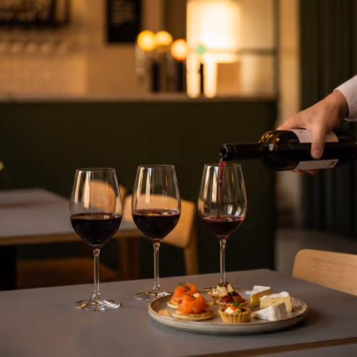 Hand pouring red wine into a glass next to two filled glasses and a plate with appetizers on a table.