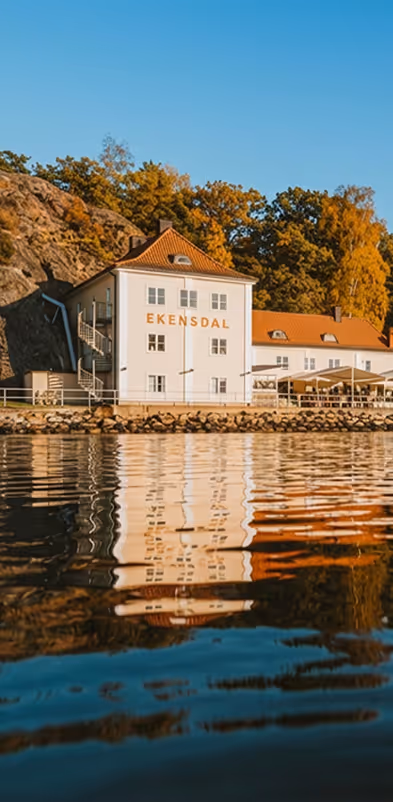 Vitt hus med rött tak och texten EKENSDAL vid vattnet med höstfärgade träd i bakgrunden.
