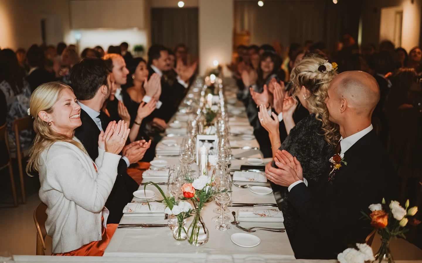 Glada gäster applåderar vid ett långbord med vita dukar och blommor under festligt evenemang.