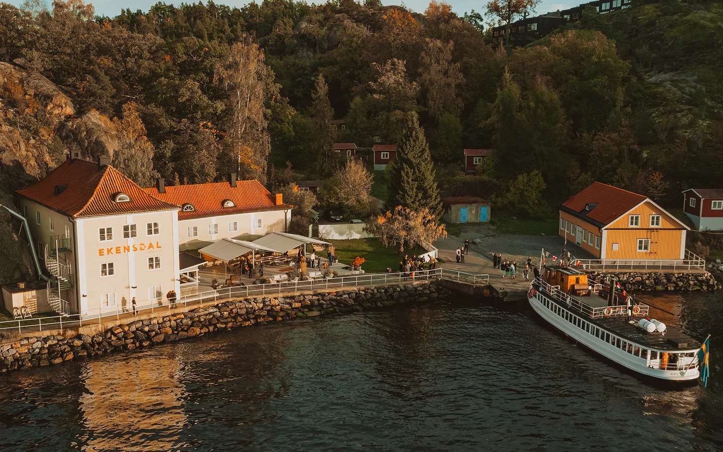 Ekensdal building by water with docked boat and autumn trees on hillside behind it.