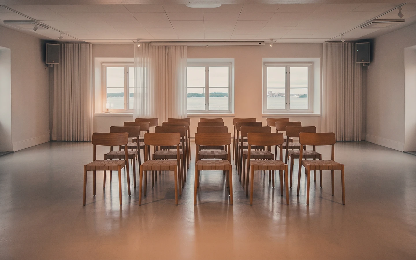 Tomt konferensrum med flera trästolar i rader och fönster med utsikt över vatten.