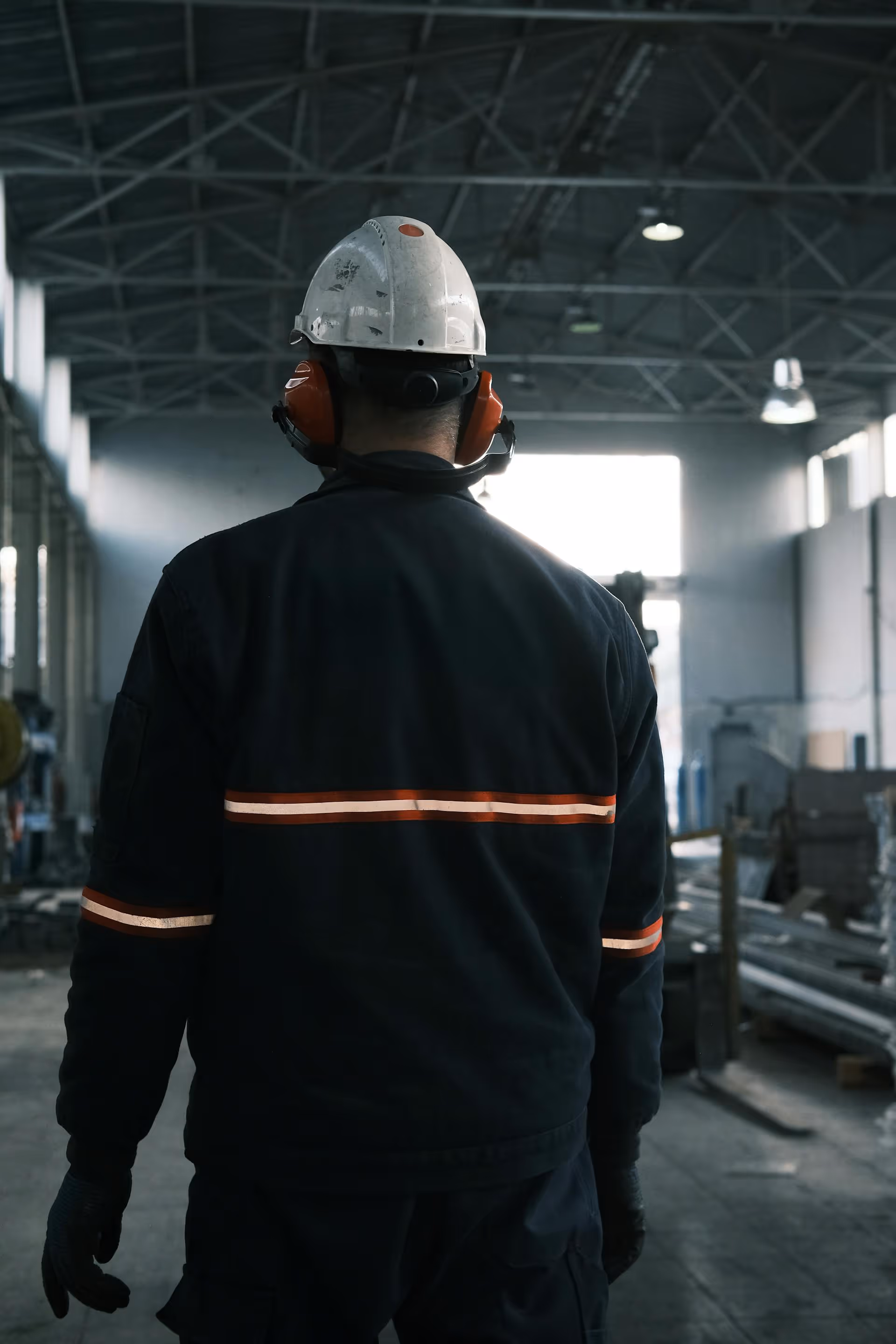 Worker in dark safety uniform with helmet and ear protection standing inside industrial warehouse.