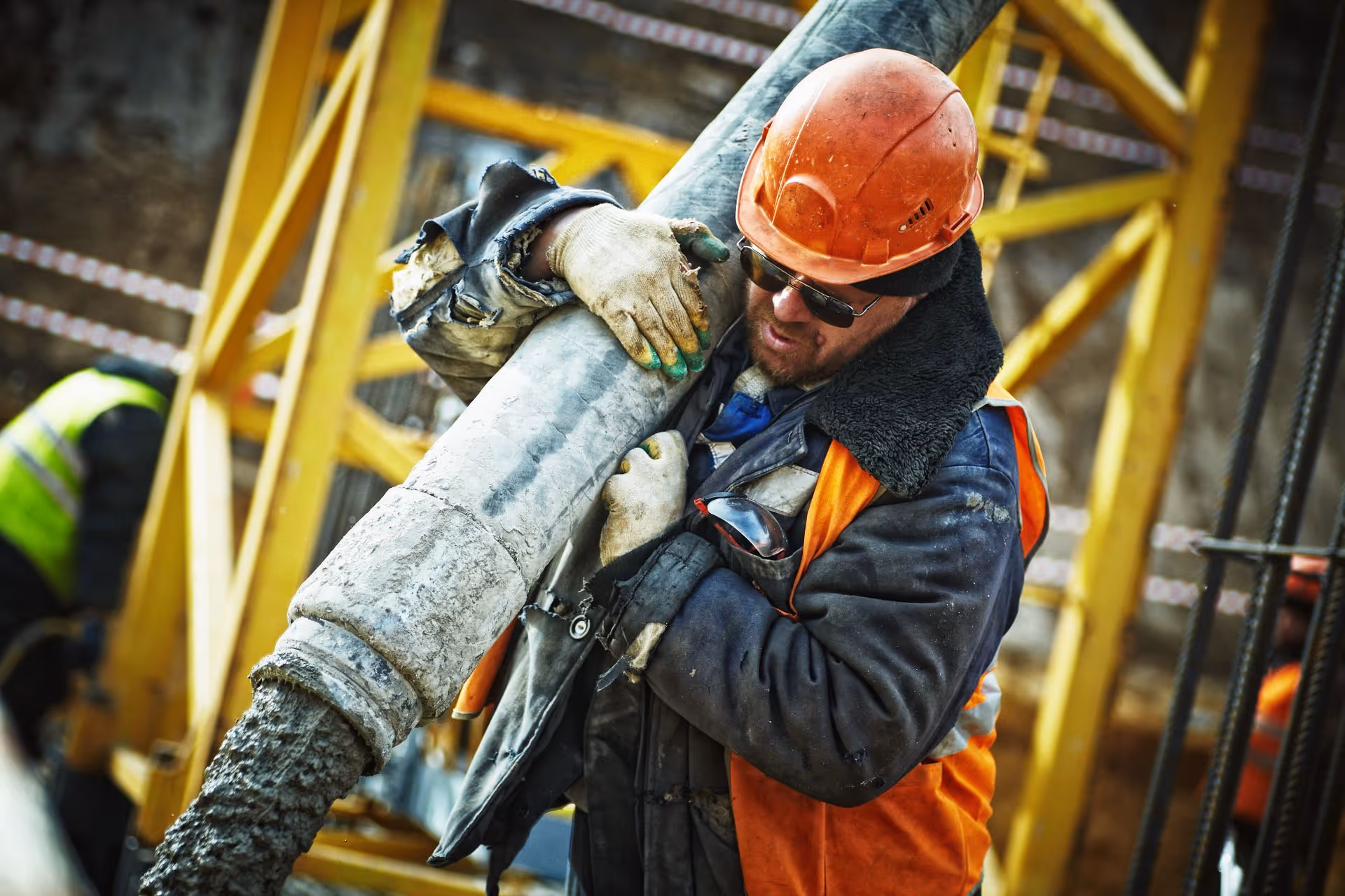 Construction worker in orange helmet and vest handling a large concrete pouring hose on site.