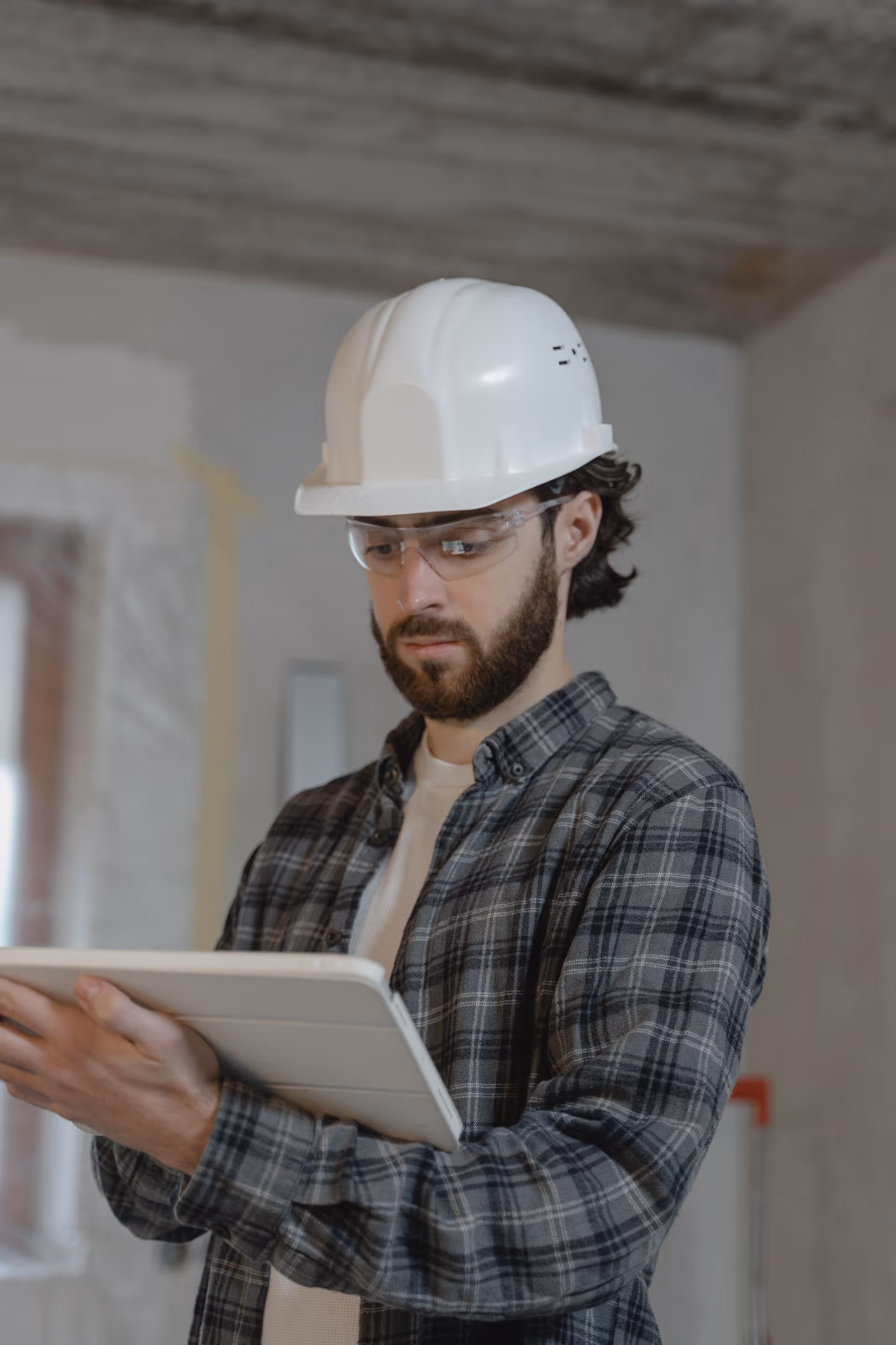 Construction worker wearing a white hard hat and safety glasses, using a tablet indoors.