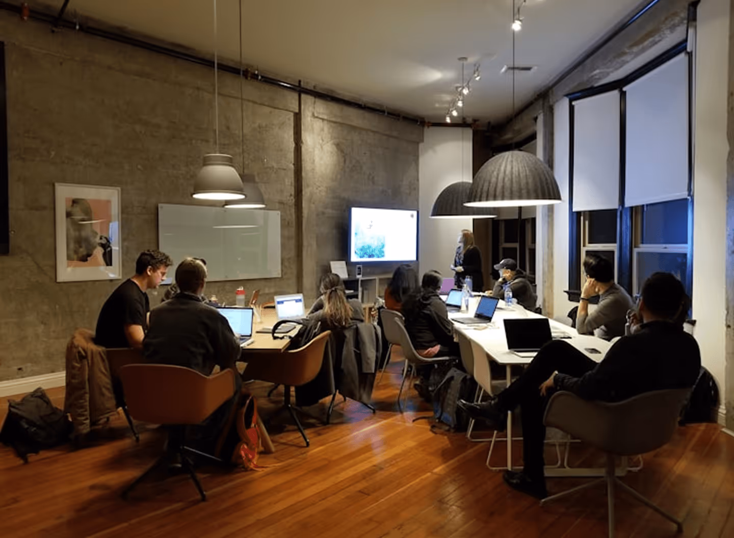 People seated around a conference table with laptops, watching a presentation on a screen in a modern office.