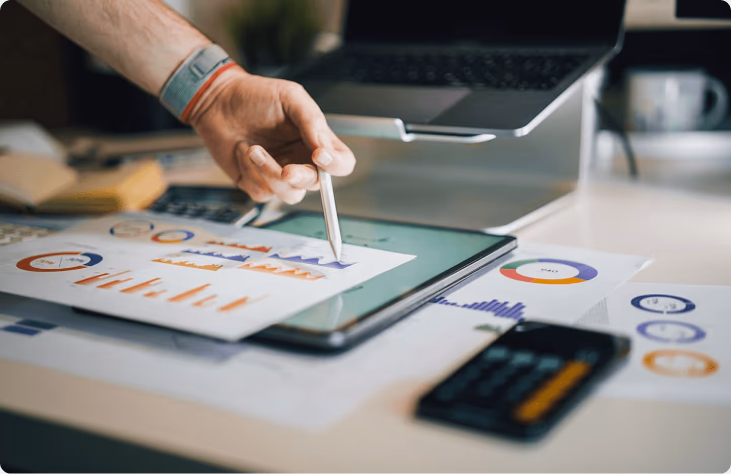 Hand holding pen pointing to charts and graphs on paper and tablet near laptop and calculator.