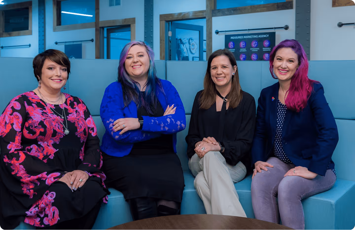 Four women sitting on a blue couch smiling in a modern office setting.