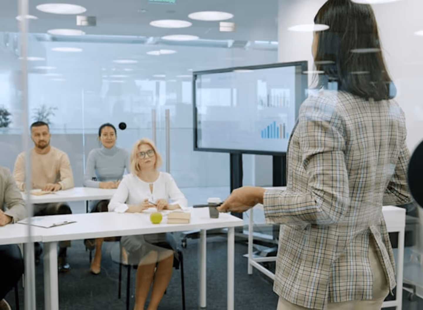 Woman in plaid blazer giving a presentation to an attentive group in a bright office.