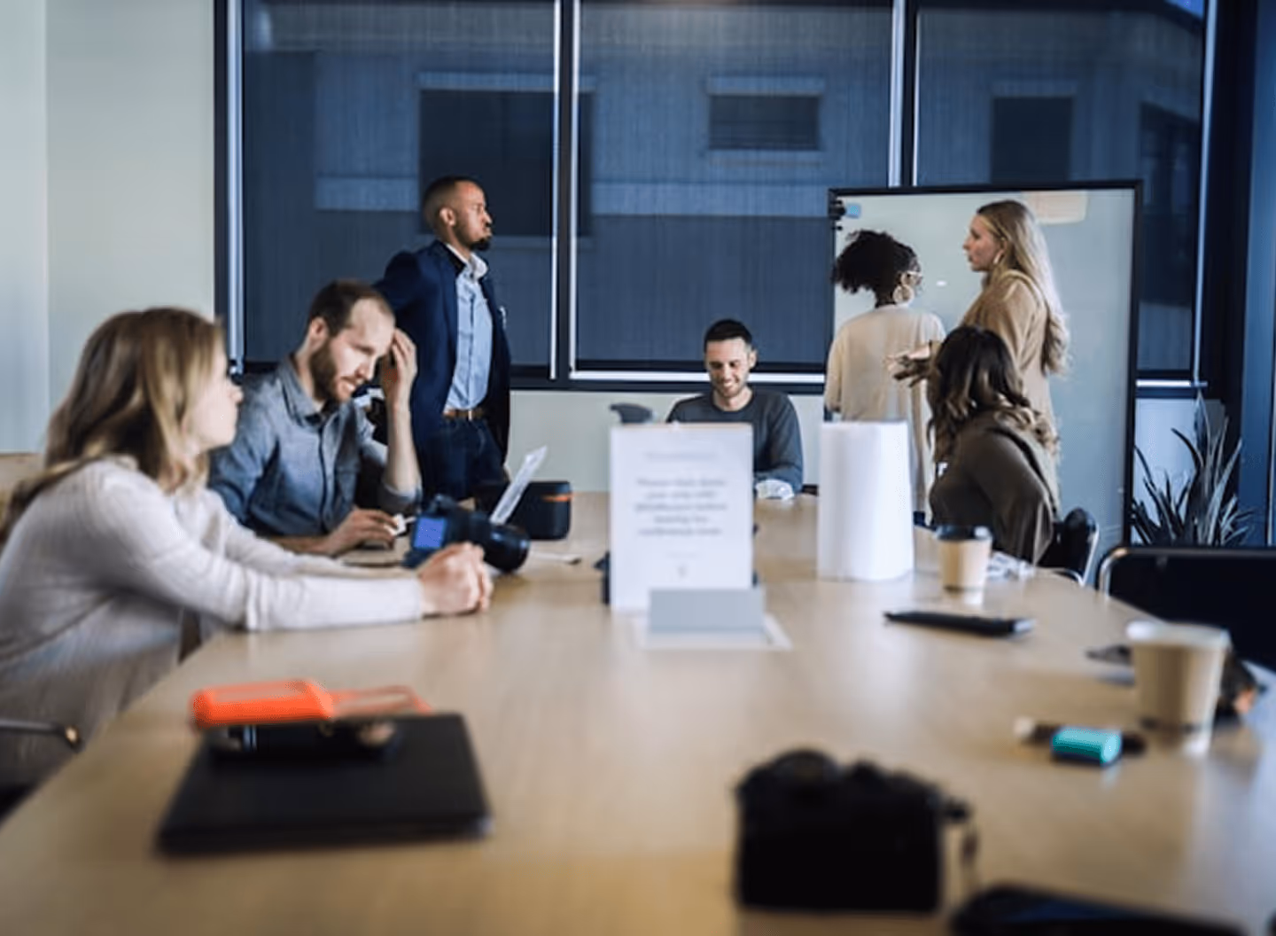 A group of diverse people in a meeting room, some seated, two women talking by a whiteboard.